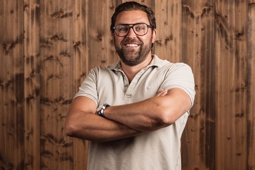 The Volgger family Smiling man wearing glasses standing in front of wooden wall