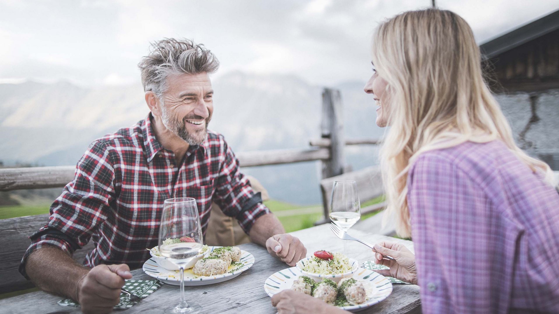 Your holiday in South Tyrol Man and woman dining outdoors on wooden bench with mountain view.
